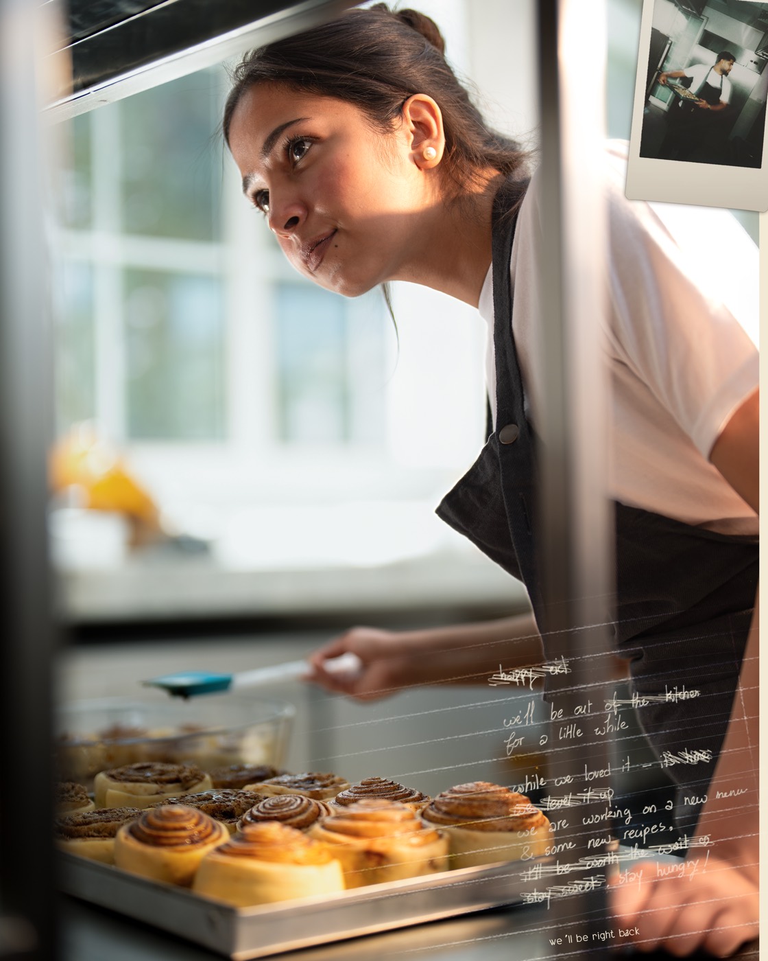 A baker leaning into a lit display case of cinnamon rolls, with a handwritten message overlaid on the glass in white.