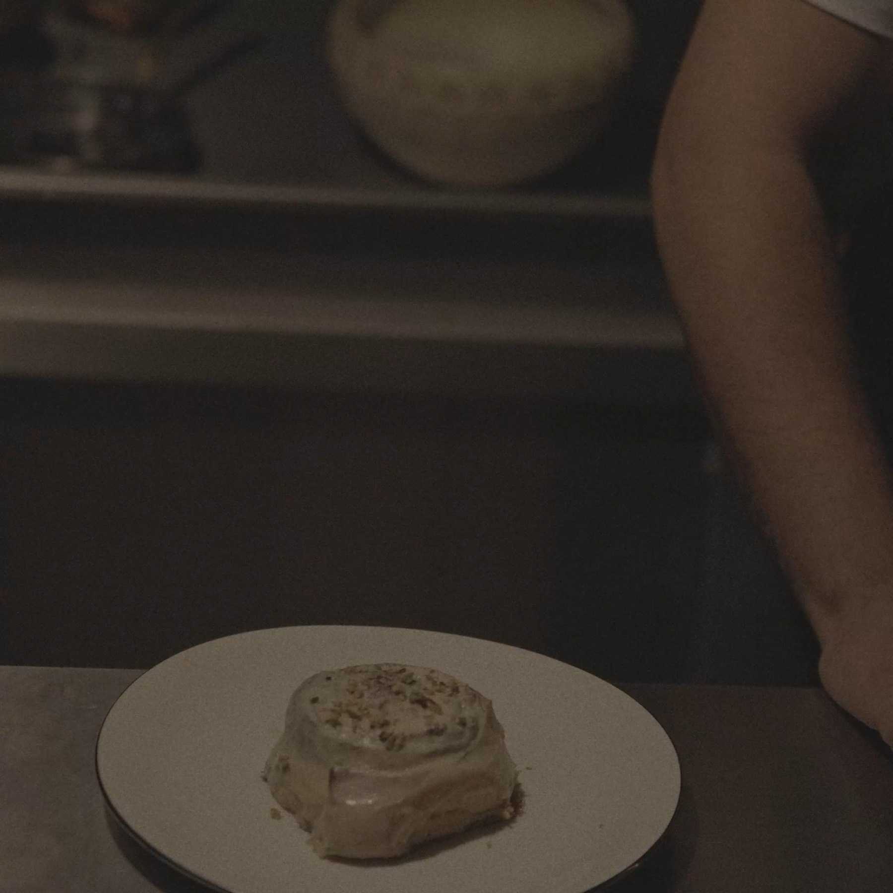 The film's opening frame. A single iced cinnamon roll sits on a pale enamel plate on a dark steel counter, lit by one soft key from the left; a baker in a white tee stands just out of focus at the right of frame, and a mixing bowl waits on the shelf behind.