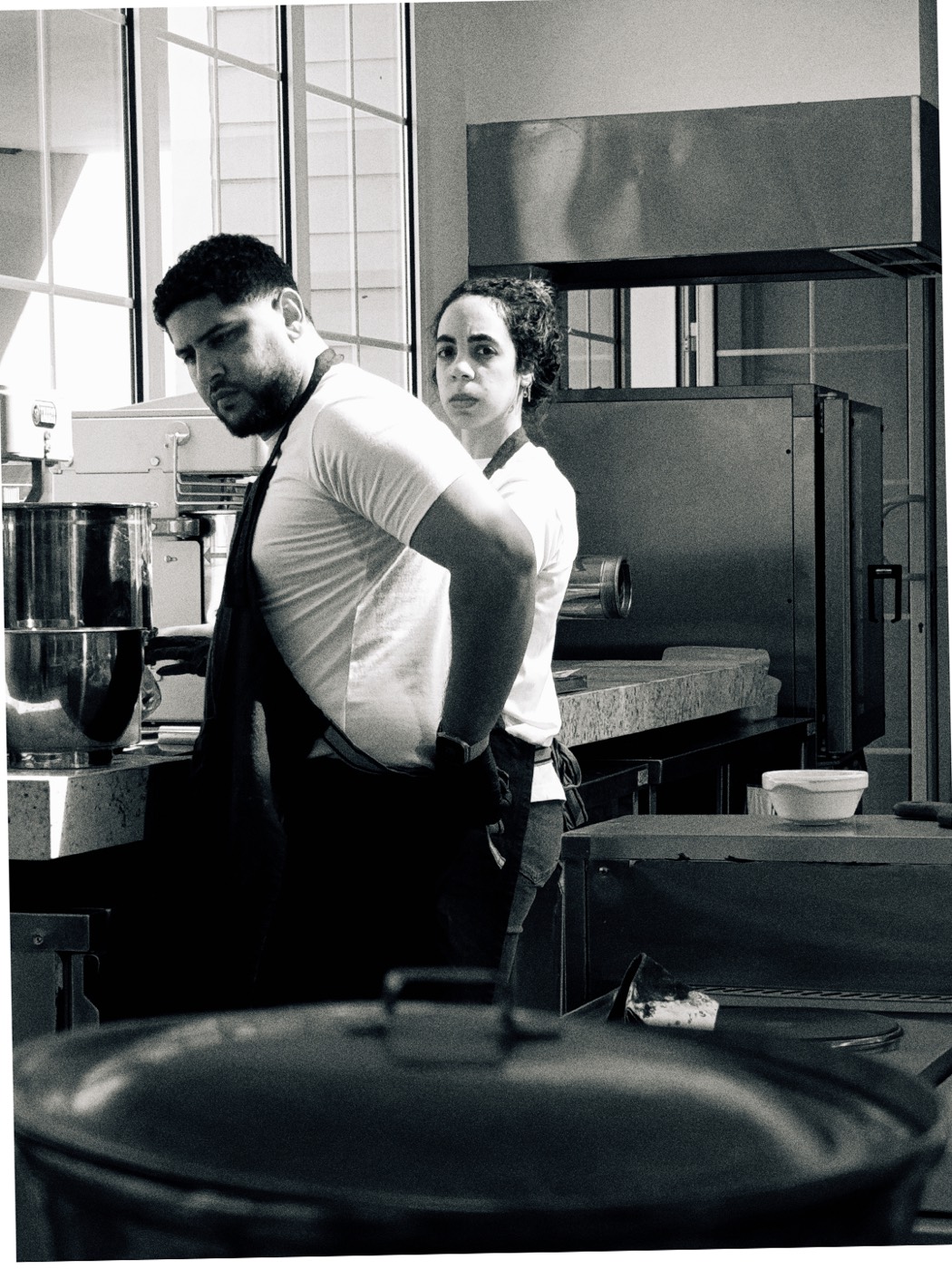 Black and white. Two bakers in a commercial kitchen, mixer and racks behind them; the one at the back has turned to look at the camera.