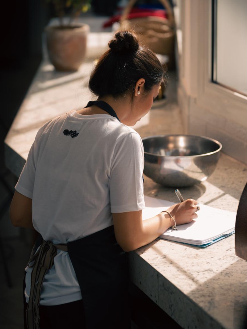 A baker leaning over a marble counter in hard morning side light, writing in an open notebook with a steel mixing bowl beside her.